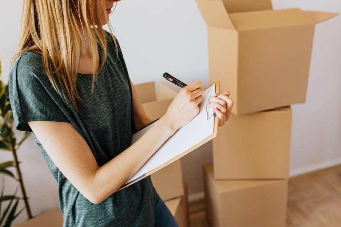 Une femme coche une liste de contrôle sur un porte-bloc au milieu de cartons, utile pour savoir comment demander l'aide mobili pass pour un déménagement professionnel.