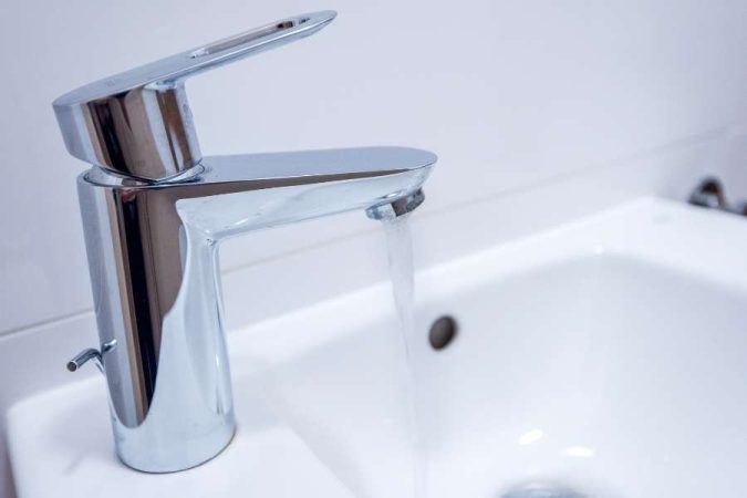 Gros plan sur un mitigeur de lavabo chromé et brillant, dont le bec verseur laisse couler un filet d'eau constant dans une vasque en céramique blanche.