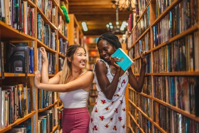 Deux amies souriantes choisissent des livres dans une grande bibliothèque sur mesure en bois.